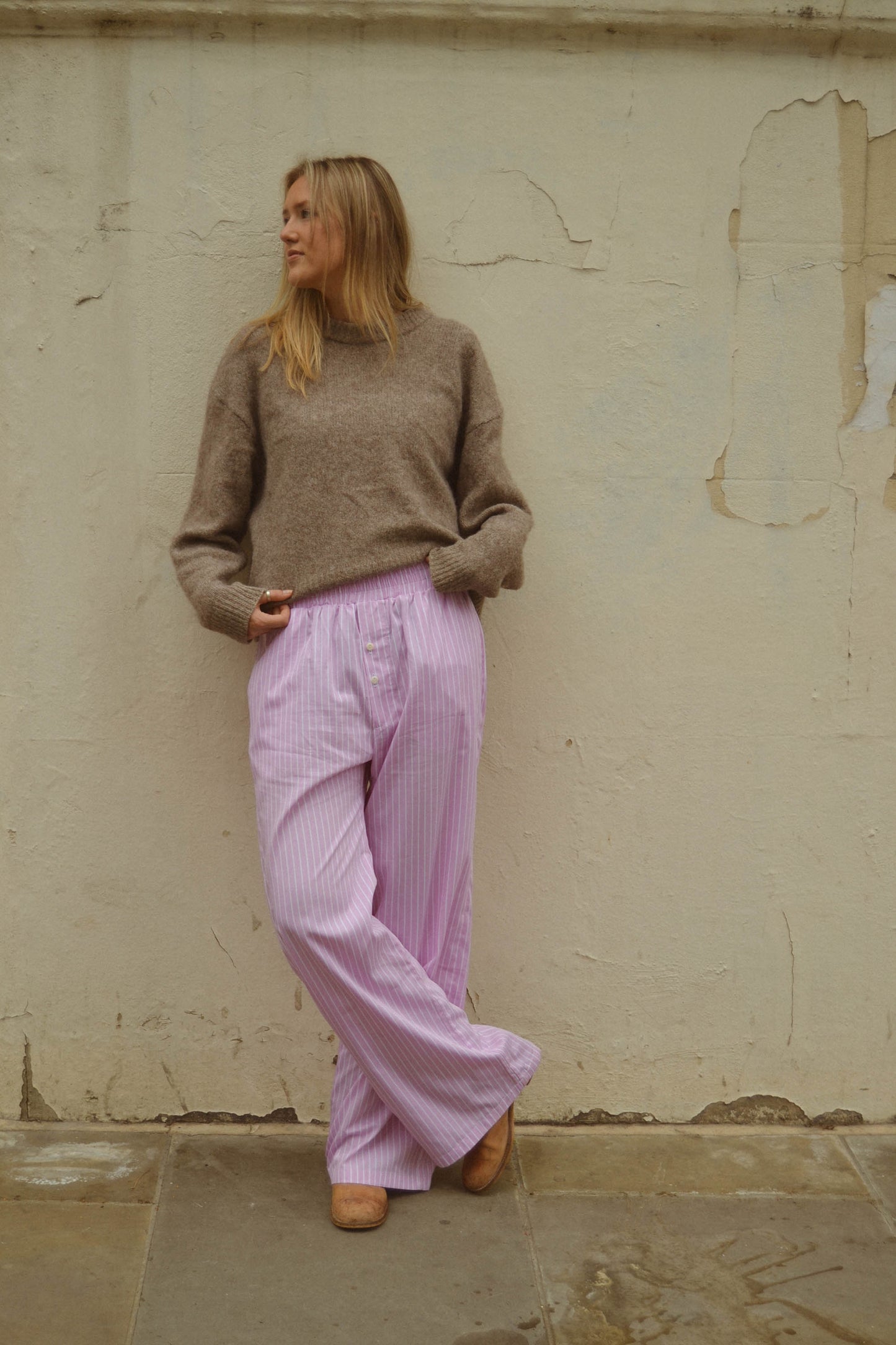 Woman wearing a brown sweater and pink pants standing against a textured wall.